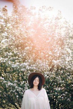 Beyaz flo ile çiçek açan ağaçta poz veren şapkalı şık boho kadın.