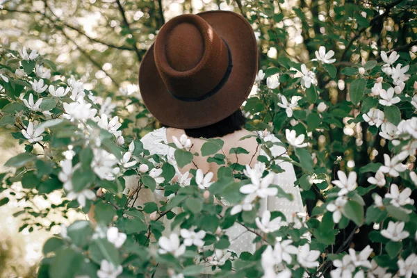Elegante mujer boho en sombrero posando en un árbol floreciente con flo blanco —  Fotos de Stock