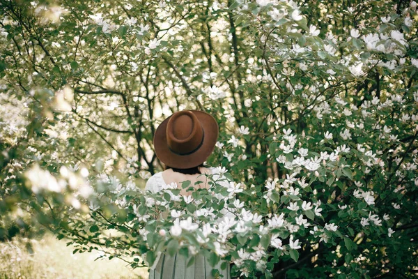Mulher boho elegante em chapéu posando na árvore florescente com flo branco — Fotografia de Stock