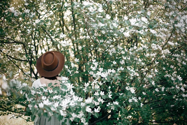 Elegante mujer boho en sombrero posando en un árbol floreciente con flo blanco — Foto de Stock