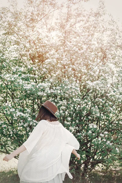 Feliz boho mujer en sombrero divirtiéndose en la luz soleada cerca de blo blanco — Foto de Stock