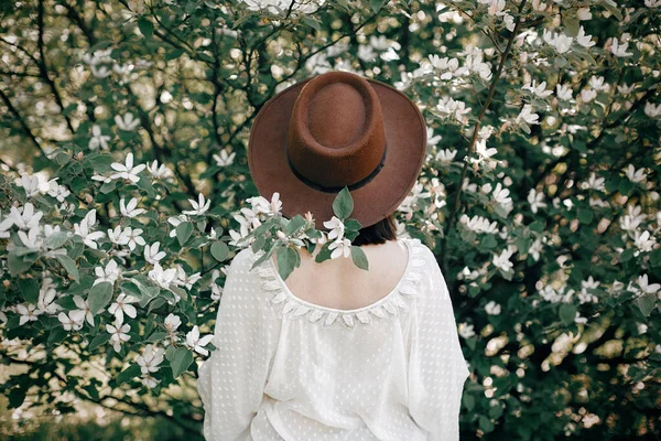 Beyaz flo ile çiçek açan ağaçta poz veren şapkalı şık boho kadın. — Stok fotoğraf