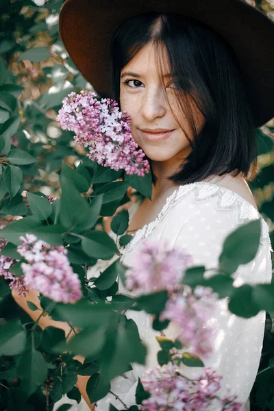 Stylish boho woman in hat posing at lilac flowers in sunny sprin — Stock Photo, Image