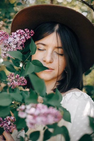 Stylish boho woman in hat posing at lilac flowers in sunny sprin — Stock Photo, Image