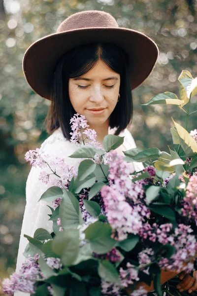 Stylish boho woman in hat holding lilac flowers bouquet in sunny — Stock Photo, Image