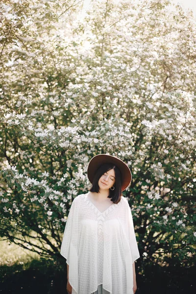 Mulher boho elegante em chapéu posando na árvore florescente com flo branco — Fotografia de Stock