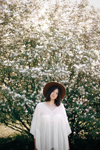 Stylish boho woman in hat posing in blooming tree with white flo — Stock Photo, Image