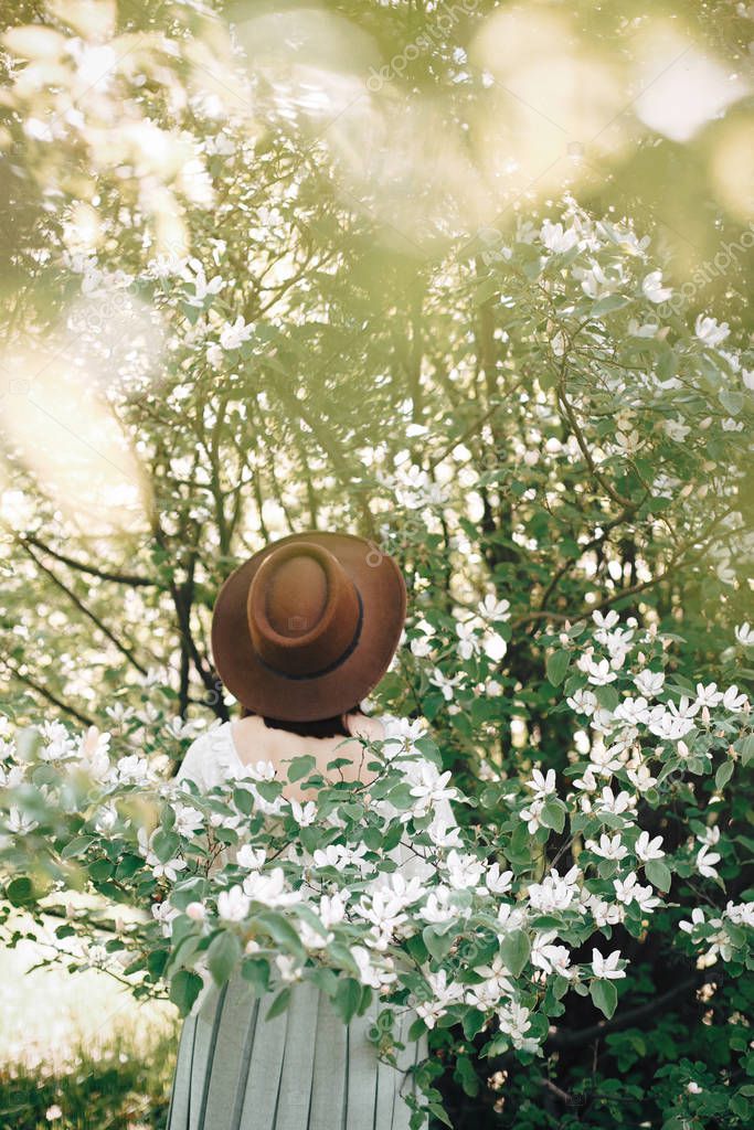 Stylish boho woman in hat posing in blooming tree with white flo