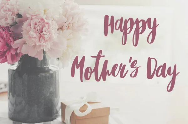 Happy mother\'s day lettering on pink and white peonies in vase and gift box with on rustic table in light, space for text. Happy mother\'s day text. Floral greeting card or poster