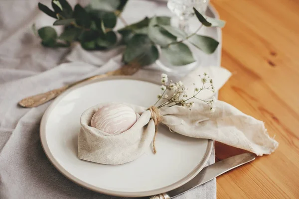 Stylish Easter brunch table setting with egg in easter bunny napkin on  table. Modern natural dyed pink easter egg on napkin with bunny ears, flowers on plate and cutlery. Easter table decorations