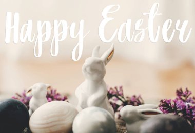 Mutlu Paskalya metinleri. Paskalya tebrik kartı. Modern Paskalya yumurtaları, beyaz tavşanlar ve keten kumaşlı leylak çiçekleri. Doğal boyanmış paskalya yumurtaları ve bahar çiçekleri. Tatil kartı