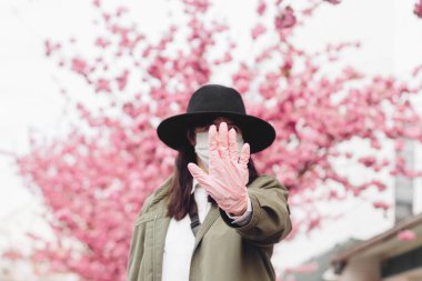 Sosyal uzaklık, uzak dur. Elindeki tıbbi eldiven virüs salgını sırasında durduğunu gösteriyor. Çiçek açan pembe ağaçların arka planında tıbbi maske takan genç bir kız. Koronavirüsü durdurun.