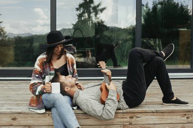 Ahşap verandada oturan şık hippi çift, dağlardaki büyük pencereli modern kulübenin arka planında ukulele ile rahatlıyorlar. Mutlu genç aile gezginleri ormanda tatilin tadını çıkarıyorlar.