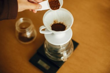 Eller filtrede öğütülmüş kahve dolduruyor. Alternatif kahve demleme v60 için hazırlanıyor. Elinde bir kaşık dolusu öğütülmüş kahveyle arka planda cam su ısıtıcısı tutan kişi.