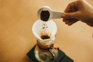 Eller filtrede öğütülmüş kahve dolduruyor. Alternatif kahve demleme v60 için hazırlanıyor. Elinde bir kaşık dolusu öğütülmüş kahveyle arka planda cam su ısıtıcısı tutan kişi.