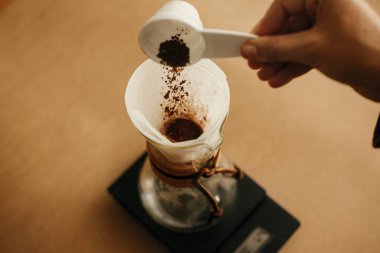 Eller filtrede öğütülmüş kahve dolduruyor. Alternatif kahve demleme v60 için hazırlanıyor. Elinde bir kaşık dolusu öğütülmüş kahveyle arka planda cam su ısıtıcısı tutan kişi.