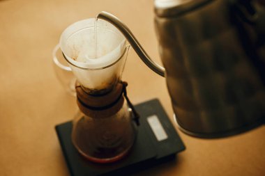 Çelik çaydanlıktan dökülen sıcak suyu filtreye dökerek ölçekte öğütülmüş kahveyle birlikte. Alternatif kahve v60 mayalı. Barista kahverengi arka planda filtre kahve yapıyor.