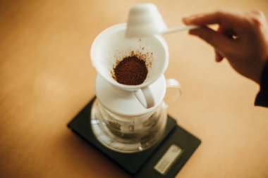 Elleri filtrede öğütülmüş kahveyi su ısıtıcısına döküyor. Alternatif kahve demleme v60 için hazırlanıyor. Kahverengi arka planda öğütülmüş kahveyle kaşık tutan kişi