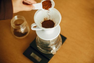 Elleri filtrede öğütülmüş kahveyi su ısıtıcısına döküyor. Alternatif kahve demleme v60 için hazırlanıyor. Kahverengi arka planda öğütülmüş kahveyle kaşık tutan kişi