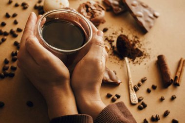 Elleri cam bardakta taze kahve ve kavrulmuş fasulye, kaşık üzerinde öğütülmüş kahve, makaron, çikolata, kahverengi arka planda tarçın. Kahve karamsar imajı