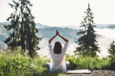 Yoga yapan ve sabah dağlarının arka planında güneşli bir ışıkta çimlerin arasında oturan genç bir kadın. Meditasyon. Sakin bir an, doğayla bağlantı. Boşluğu kopyala