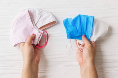 Yeniden kullanılabilir yüz maskeleri ve beyaz ahşap arka planda tek kullanımlık tıbbi maskeler tutan eller. Virüs salgını sırasında sıfır atık yaşam tarzı. Tek kullanımlık maske ve el yapımı tekstil maskesi