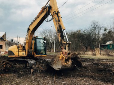 Kazıcı, kırsaldaki ağaçların kökünü kazıyor. Buldozer eski ağaçları, kökleri ve dalları toprak ve çöplerle temizliyor. Kazma makineleri. Bahçe işleri
