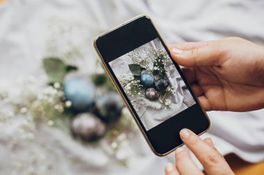 Telefon tutan ve şık paskalya yumurtalarının fotoğrafını çeken eller beyaz bahar çiçekleri ile kırsal masada yatıyordu. Kırsal tekstil arka planında doğal renkli paskalya yumurtaları. Metin için boşluk