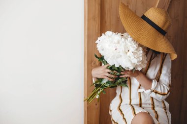 Uluslararası Kadın Günleri. Beyaz şakayıklı buketi taşralı ahşap arka planda tutan şapkalı şık bohem kadın. Mesaj için yer var. Şakayıklarla poz veren hippi kız. Anneler Günün kutlu olsun..