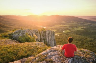 Kayalık bir tepe üzerinde oturan adam ve güneşin batışını