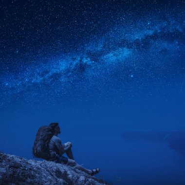 Sırt çantalı bir yürüyüşçü denizin üzerindeki bir uçurumda oturuyor ve yıldızlı bir gecede Samanyolu 'nun tadını çıkarıyor..