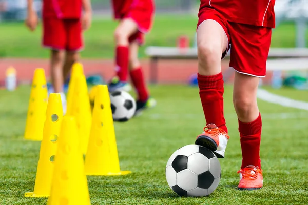 Детские футбольные тренировки. Молодой спортсмен с футбольным мячом на поле. Ребенок в форме для красного футбола. Тренировки по дриблингу на спортивном травяном поле — стоковое фото