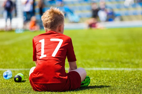 Jovem jogador de futebol. Atleta Juvenil no Red Football Sportswear. Menino sentado sozinho no campo de futebol e assistindo torneio de futebol juvenil . — Fotografia de Stock