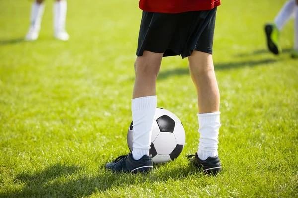 Закройте ноги и ноги футболиста в белых носках и черных туфлях, играя с типичным мячом, стоящим на зеленой траве на открытом воздухе. Ребёнок играет в футбол — стоковое фото