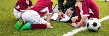 Futbol takımının koçu. Gençlik sporları koçu taktik tahtası kullanıyor. Antrenör maç stratejisini açıklıyor. Spor futbol eğitimi. Üç çaylak oyuncu çim sahada koçla oturuyor.