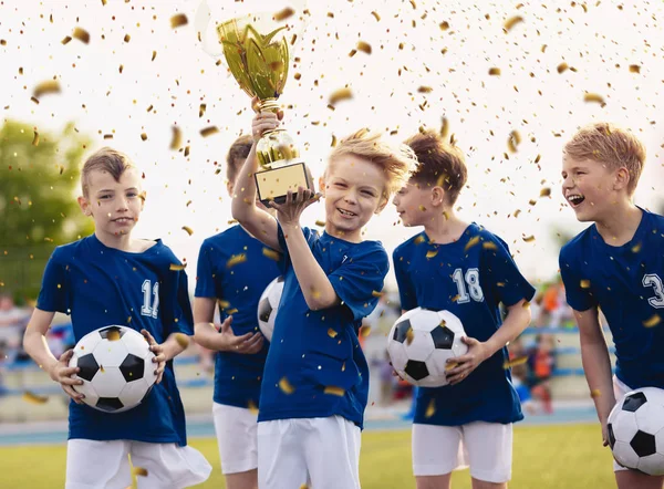 トーナメント決勝でサッカーの成功を祝う小学生スポーツチームの幸せな子供たち。少年たちは笑顔とトロフィーの儀式に黄金のカップを上げ — ストック写真