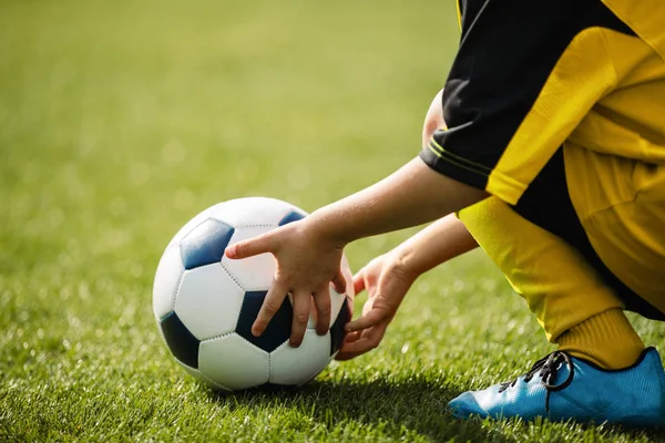 Αγνώριστο παιδί που παίζει με μπάλα ποδοσφαίρου στο γήπεδο. Μικρό παιδί που κρατάει και τα δύο χέρια μπάλα ποδοσφαίρου. Νεαρός ποδοσφαιριστής με μπλε αθλητικά παπούτσια — Φωτογραφία Αρχείου
