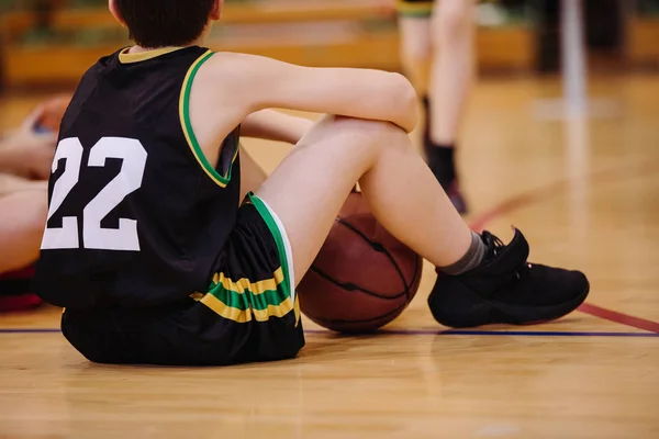 Младший баскетболист сидит на полу на баскетбольной площадке — стоковое фото