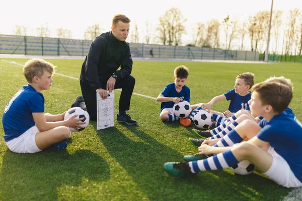 Een groep jonge jongens zit op Sports Grass Field heksenschool coach. Kinderen luisteren naar de tactiek van de coach. De jonge coach legt de voetbaltactiek uit. Coaching Jeugd in Sport — Stockfoto