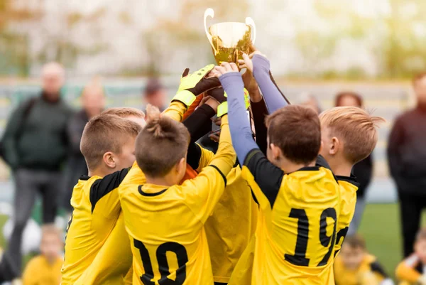 İlkokul spor takımında sarı tişörtlü mutlu çocuklar turnuva finalinde futbol başarısını kutluyorlar. Spor takımı üyeleri altın kupa kupasını yükseltiyor — Stok fotoğraf