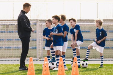 Genç koç, çocukların futbol takımına antrenman kurallarını anlatıyor. Futbol takımındaki çocuklar koçu dinliyor. Çocuklar futbol antrenmanında. Açık havada beden eğitimi dersi.