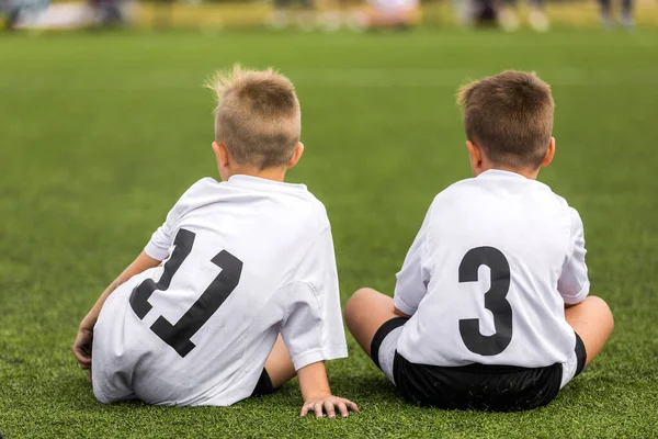 Two boys in sports uniforms sitting on grass pitch. Kids playing team sports