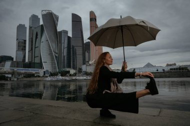 Kötü hava, insan ve konsept manzarası - Vrikshasana 'da şemsiyeli genç gülen iş kadını - Moskva Nehri' nin setinde Moskova İş Merkezi 'ne karşı ağaç duruşu