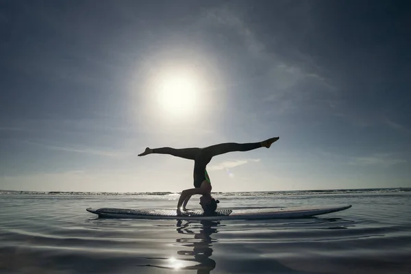 瑜伽头架变化海背景 一个女人在海里用体操纵向绳在长板上表演床头倒立 — 图库照片