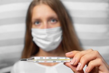 Gripten etkilenmiş tıbbi maske takan genç kadın termometrede yüksek sıcaklıkta yattığını gösteriyor. Zatürree salgını, Coronavirüs salgını kavramı,