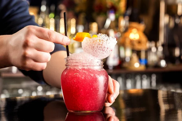 Bartender is decorating red cocktail in jar with frappe at bar counter background.