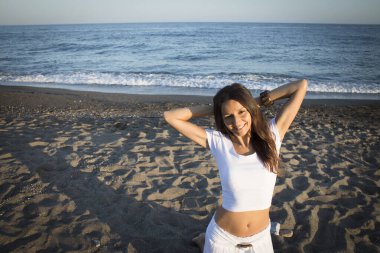 Plajda beyaz tişörtlü ve kolları boynunda olan bir kız. Çok mutluyum.