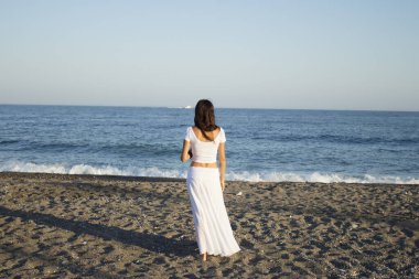 Plaja arkası dönük beyaz elbiseli bir kız. Sakin sahne