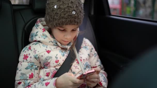 Liten flicka i vinter hatt och rock i bilen spelar telefon. — Stockvideo