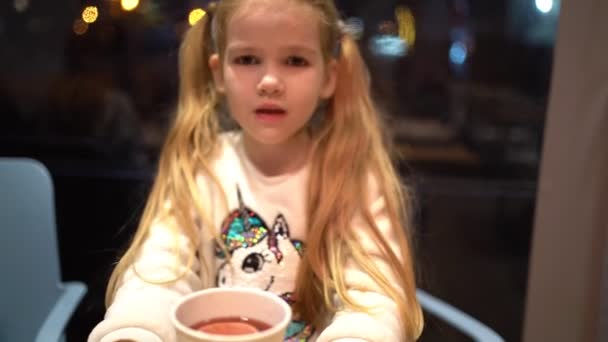 Маленькая блондинка с хвостами пьет горячий чай вечером в кафе . — стоковое видео